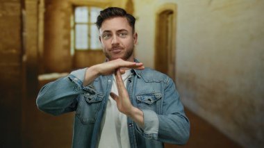Kot ceketli genç adam eski bir üniversite binasında sıcak aydınlatma, rahatlama ve duraklamayı sembolize eden bir jest yapıyor..