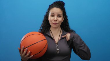 İspanyol kadın antrenör elinde bir basketbol topu tutarken boynunda bir ıslıkla canlı mavi bir arka plan tutuyor..