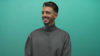 Stüdyoda yeşil panelde küpe takılı gri balıkçı yaka giyen, geniş bir gülümseme ve sakal gösteren bir adam; sıcaklık.