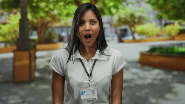 Güneşli bir parkta gülümseyen bir kadın, etrafı yemyeşil ağaçlarla çevrili bir kimlik kartı ile açık havada neşeli ve profesyonel bir atmosfer yaratıyor..