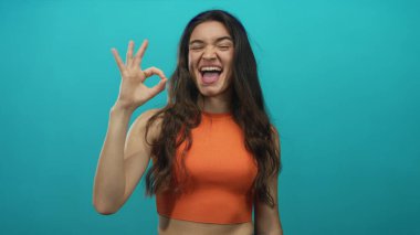 Turuncu bluz giyen bir kadın açık bir şekilde gülümserken deniz mavisi atölyesinde el işareti gösteriyor; iyimserlik..