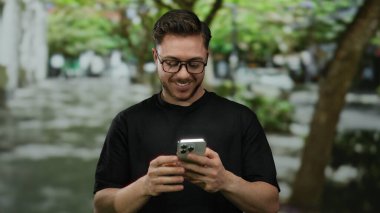Sakallı ve gözlüklü genç adam, şehir caddesinde akıllı telefona bakarken gülümsüyor, arka planda ağaçlarla şehir merkezinin tadını çıkarıyor..