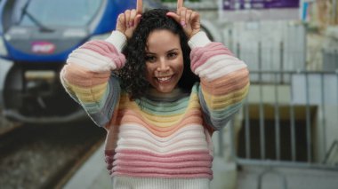 Renkli kazaklı genç İspanyol kadın açık bir günde arka planda canlı bir trenle açık hava tren istasyonunda şakacı boynuzlu jest yapıyor..