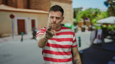 Kırmızı çizgili gömlekli genç adam, şehir sokağında kolunda dövmeleri olan etkileyici hareketler yapıyor.