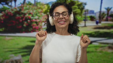 Güneşli bir parkta kulaklıkla müzik dinleyen bir kadın, yemyeşil ve çiçeklerle çevrili, gözlük takıyor ve beyaz gömlek giyiyor, gülümsüyor ve rahatlıyor..