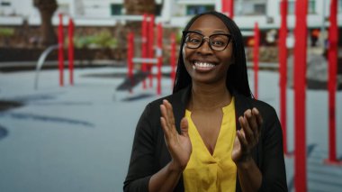 Dışarıdaki spor salonunda kırmızı ekipmanlarla gülümseyen bir kadın. Gözlüklü, sarı tişörtlü, gündelik tarzda ve güneşli gökyüzünün altında neşeli bir ifade. Mutluluk ve sıcaklığı yansıtıyor..