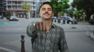 Şablon gömlekli genç İspanyol adam şehrin arka planında güneşli bir gökyüzünün altında yumruğuyla güvenle gülümsüyor..