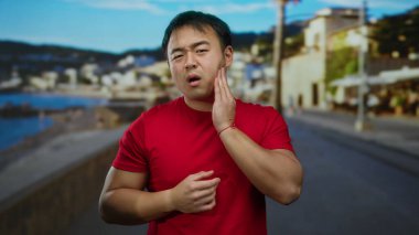 Kırmızı gömlekli genç adam deniz kenarında bir gezinti güvertesinde dikilirken diş ağrısı çekiyor. Arka planda mavi gökyüzünün altında bulanık bir sahil köyü var..