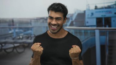 Genç Hispanik adam geniş bir gülümseme ve deniz kenarı binasında yumruklarını sıkar; heyecan sevinç coşkusu coşkusu.