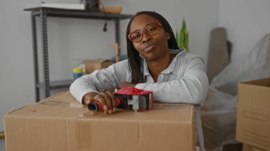 Yeni evdeki kutuları bant kutusuyla dolduran kadın paketlenmemiş eşyalarla çevrili. Yorgun görünüyor ama günlük kıyafetlerle kararlı..