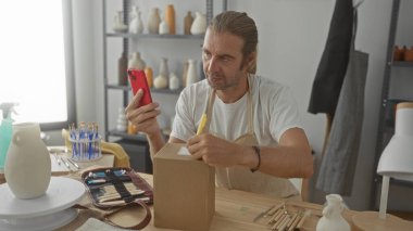 Önlük takan sarışın adam akıllı telefonu tutuyor ve dağınık bir sanat stüdyosundaki karton bir kutuya yazıyor. İçinde çömlek rafları ve aletler var..