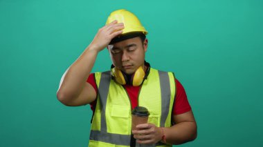Asyalı bir adam inşaat malzemeleri giyiyor ve elinde kahve fincanı var. Yeşil arka planda duruyor. Yorgun görünüyor..