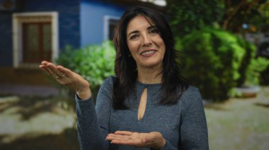 Orta yaşlı İspanyol kadın, yemyeşil park yeşilliklerinin ortasında binanın önünde avuçlarını açık tutuyor..