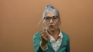 Gri saçlı ve gözlüklü yaşlı kadın, yeşil bir kazak giyen, izole edilmiş kahverengi bir arka plana sıkıca tutunarak öfkesini ifade ediyor..