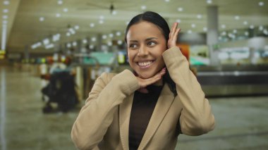 Havaalanı terminalinde gülümseyen kadın çerçeve hareketleri yapıyor, seyahat ve gençliği temsil ediyor; arka plan gösterimi meşgul, modern havaalanı ambiyansı.