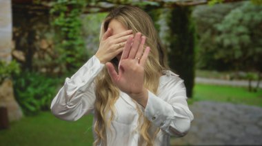 Parkta yeşil yapraklı bir kadın, açık beyaz gömlekli, yemyeşil ve doğal ortamda, yüzü elle kaplanmış bir şekilde duygusal tepki veriyor..
