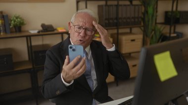 Ofiste bilgisayar başında çalışan kıdemli bir adam, akıllı telefonunu kontrol ediyor, etrafı kitaplar, bitkiler ve kırtasiye malzemeleriyle çevrili, profesyonel bir iş yeri düzeneği kuruyor..