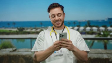 Deniz kenarındaki gezinti güvertesinde akıllı telefon kullanırken gülümseyen sakallı genç bir doktor, steteskop tutuyor, çarpıcı mavi deniz zeminine karşı modern sağlık hizmetleri iletişimi sergiliyor..