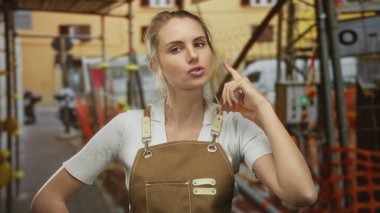 Önlüklü sarışın kadın, parlak arka plan renkleri ve endüstriyel unsurları olan hareketli bir inşaat alanında düşünceli bir şekilde kafa yoruyor..