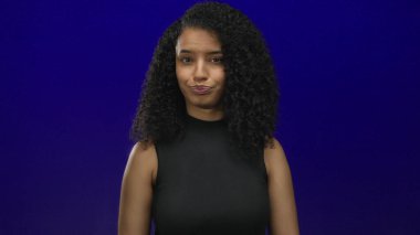 Genç Latin bir kadın, mavi bir duvara karşı gözünün üstüne işaret ve orta parmağını koyuyor..