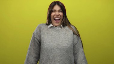 Parlak sarı bir duvarın önünde sevinçle gülen bir kadın, parlak arkaplana karşı rahat gri bir kazak giyiyor, mutluluk ve enerji saçıyor..