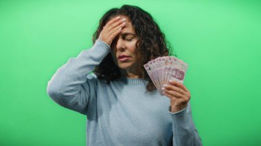 Orta yaşlı bir kadın, yeşil bir arka plan duvarına karşı Meksika pezosu tutuyor. Finansal kaygıları ve para yönetimi tasvir ediyor..