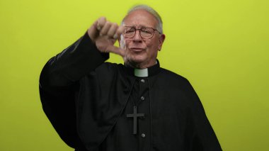 Siyah cüppeli kıdemli rahip, parlak sarı bir arka plana baş parmak işareti yaparak ciddi bir ifadeyle onaylamadığını iletiyor..