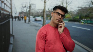 Kırmızı kazaklı genç adam arka planda kırmızı çift katlı otobüslerle şehir caddesinde telefonla konuşuyor.