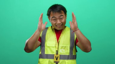 Sarı yelekli genç adam el işaretleriyle yeşil arka planı imzalıyor neşeli bir gülümsemeyle ifadesini gösteriyor.
