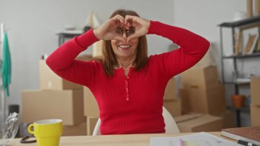 Kırmızı kazaklı İspanyol kadın yeni dairesinin içinde elleri ile kalp oluşturuyor. Kutularla çevrili, rahat bir oturma odasında sevinç ve heyecan ifade ediyor..