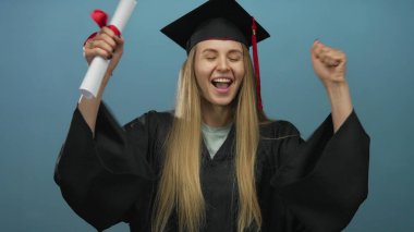 Mezuniyeti kutlayan kadın diplomasını mavi arka planda tutarken neşeli bir ifadeyle akademik önlük ve kep takıyor..