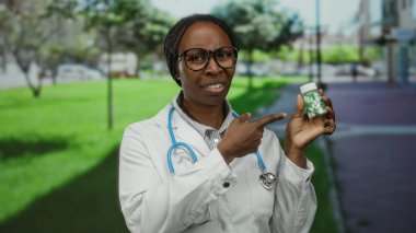 Parkta gözlüklü ve steteskoplu beyaz önlüklü bir kadın yeşil ve açık hava ile çevrili bir ilaç şişesini tutuyor ve işaret ediyor..