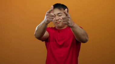 Kırmızı gömlekli genç adam turuncu arka plana parmak tabancasıyla el hareketi yapıyor, tenha bir ortamda güven ve neşe taşıyor..