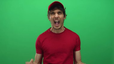 Kırmızı şapkalı ve gömlekli bir adam açık ağızla bağırıyor ve stüdyoda yeşil arka plana el kaldırıyor. Öfke öfkesinin öfkesine yenik düşüyor..