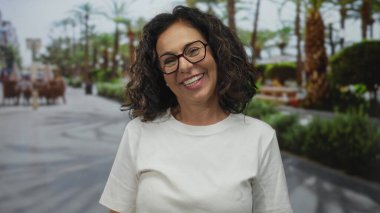 Orta yaşlı kıvırcık saçlı ve gözlüklü İspanyol kadın sokakta neşeli bir sırıtışla ağzını açar..