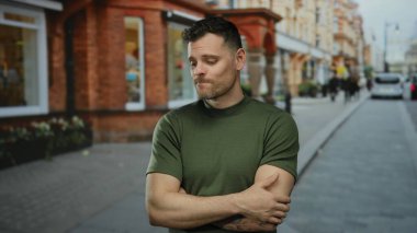 Yeşil gömlekli genç adam bir şehir caddesinde düşünceli bir şekilde duruyor, şehir manzarasına derinlik katıyor, modern hayatın ruh halini ve karakterini yansıtıyor..