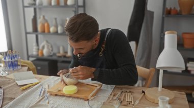 Donanma önlüklü İspanyol adam, stüdyo atölyesinde ahşap masa üzerine metal oyma aleti ile sert kilden kase oyuyor..
