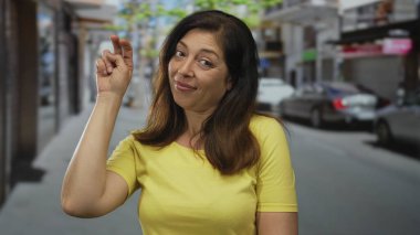 Orta yaşlı İspanyol kadın, park halindeki arabaların yanında parmağını şıklatırken elini kaldırıyor..