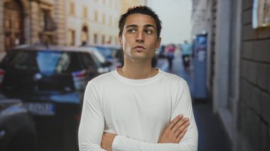 Bir şehir sokağında çapraz kolları olan İspanyol genç adam, arka planda bulanık arabaları ve yayaları ile hareketli bir şehir arka planında şehir yaşamını yansıtıyor..