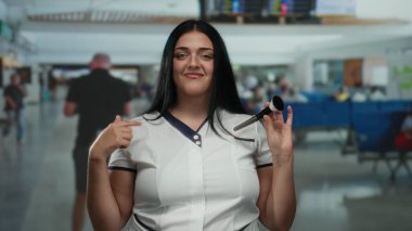 Havaalanı terminalinde makyajını hazırlarken gülümseyen bir kadın, kapalı bir alanda kendine güvenen, kıvrımlı, artı boyutta bir varlığı ön plana çıkarıyor..