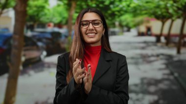 Açık bir sokakta, yeşil yapraklı bir tepe örtüsünün altında canlı bir şehir ortamında gülümseyen gözlük takan bir kadın..