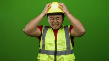 Sarı kasklı ve yelekli bir adam, parlak yeşil arka planda yüzünü buruşturuyor, elleri başının üstünde stres yapıyor..