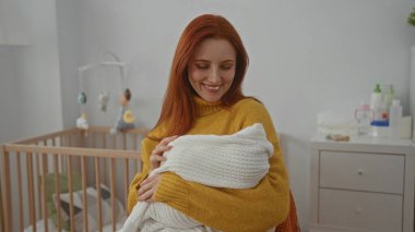 Kadın kucağında bebek beşiği, arka planda görünür bir beşikle kucağında gülümseyerek sıcak ve şefkat dolu sakin bir iç mekan sahnesi sergiliyor..