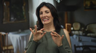 Kadın iki elini de kaldırıyor ve restoranda bir masada otururken sırıtıyor arkasında sandalyeler ve perdelerle; neşe.