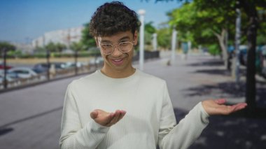 Yuvarlak gözlüklü ve beyaz bluzlu bir adam gülerken ağaçlık şehir sokağında avuçlarını açık tutar; belirsizlik..