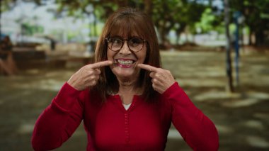 Kıdemli İspanyol kadın, güneşli bir açık hava parkı ortamında gülümseyip dişlerini gösteriyor. Keyif ve sıcaklık saçıyor. Günlük tarzıyla ve doğal güzelliğiyle..