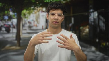 Gözlüklü adam, şehir sokağında ağaçlarla kaplı beyaz gömleğini çekiştiriyor ve yaz güneşinin altında park edilmiş arabaları çekiyor; rahatsızlık verici..