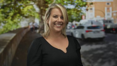 Şehir caddesinde ağaçlarla ve park edilmiş arabalarla çıplak omuzlarıyla gülümseyen bir kadın, siyah bir bluz giyiyor. Mutluluk..