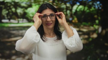 Beyaz bluz giyen orta yaşlı bir kadın yeşil park ortamında gözlük ve parmaklarını düzeltiyor; kendine güven huzuru.