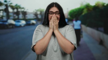 Bir şehir caddesinde elleri kapalı gülümseyen bir kadın güneşli gökyüzünün altında neşe saçıyor..